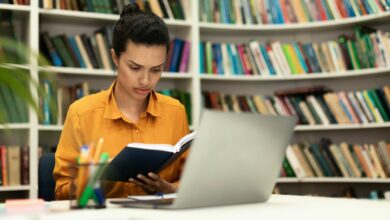 Concentrated mixed race woman reading book, sitting in library with laptop pc and study materials