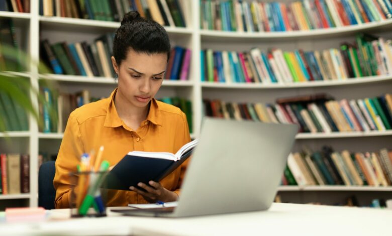 Concentrated mixed race woman reading book, sitting in library with laptop pc and study materials