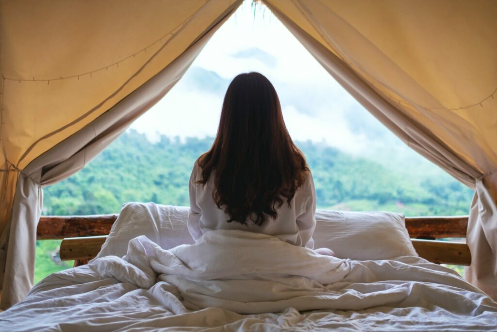 a woman sitting on the bed and looking at a beautiful nature view outside the tent
