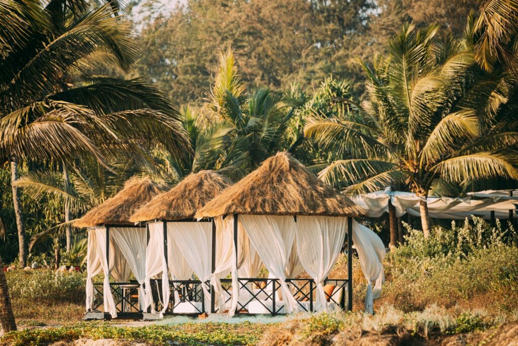 Goa, India. Gazebo Tents With Strawing Roof For Tourists On Beach With Tables And Sunbeds Inside